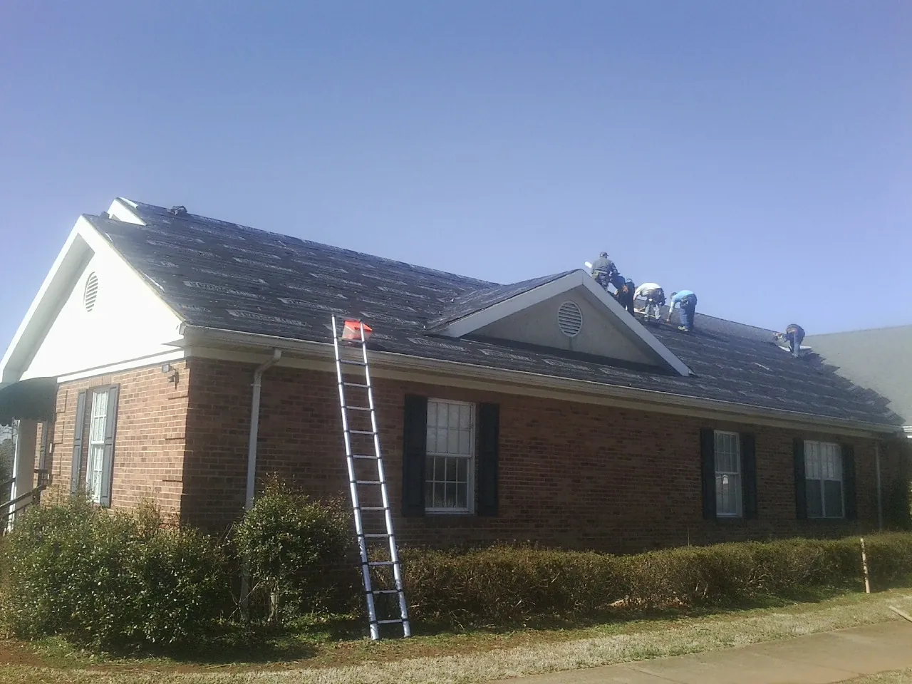 Crew tearing shingles off a brick ranch roof with a ladder set — Metal Roof Repair work by our crew in Lawsonville