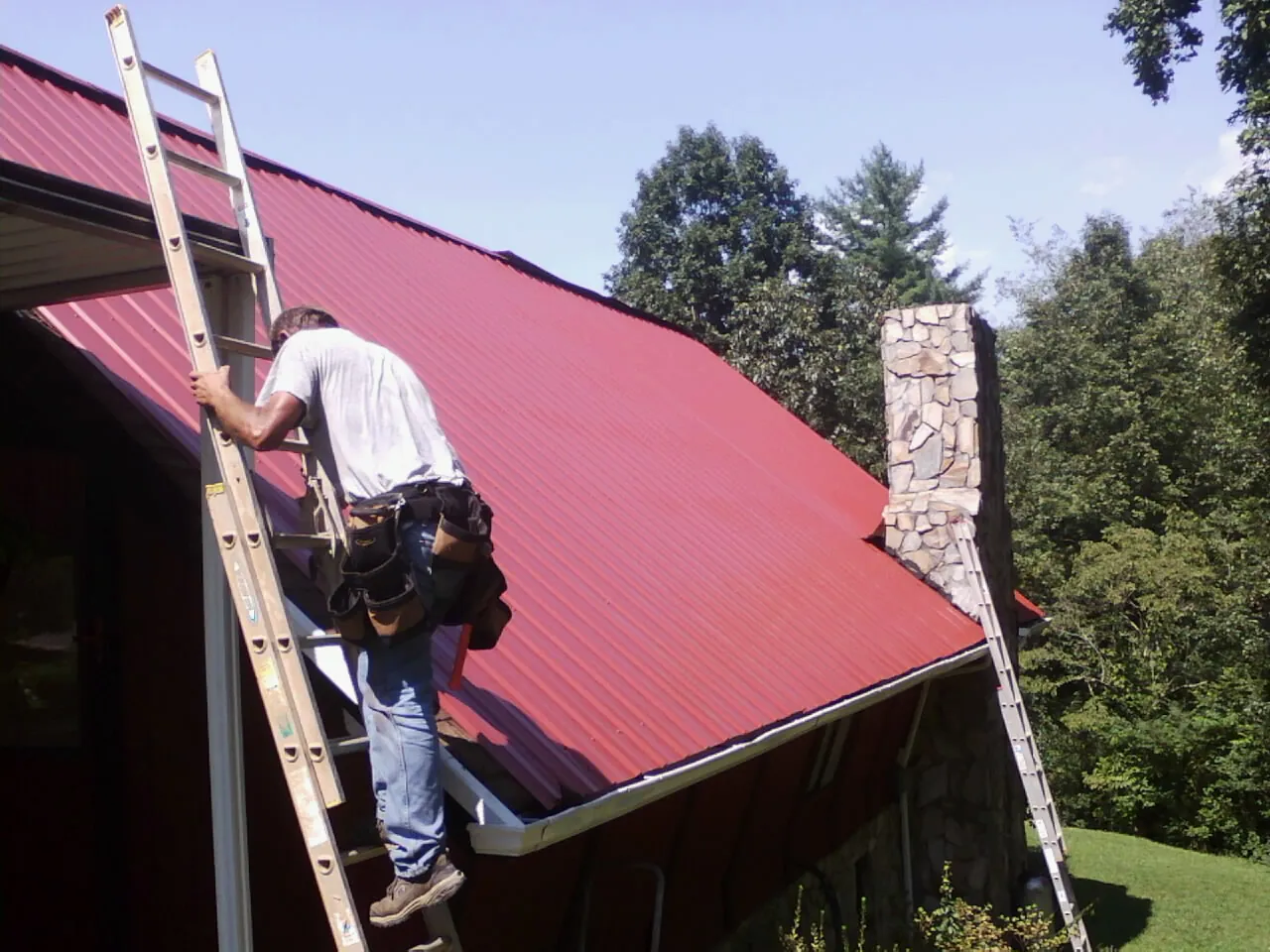 Roofer on a ladder beside a red metal roof and stone chimney — Soffit & Fascia Repair by Roofing Brothers in Lawsonville